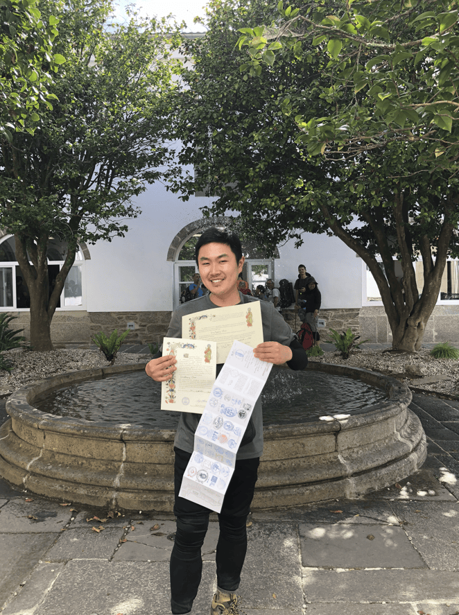 Ben (Sung Kyeong) holding his official Compostela certificate of completion for the Camino de Santiago, smiling broadly in Santiago de Compostela.