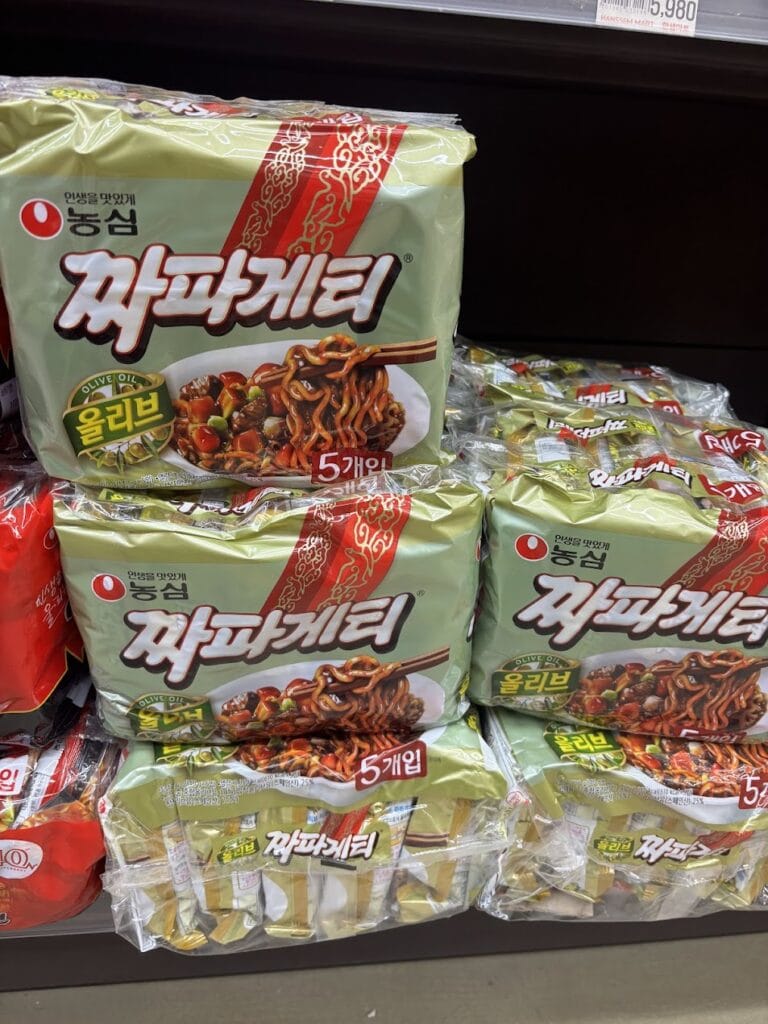 Shelves at a supermarket stocked with Nongshim Chapagetti (Korean black bean instant noodles) in its iconic olive-green and gold packaging.
