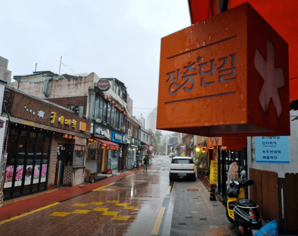 rainy day scene of the famous Jangchung-dan Jokbal Street in Seoul, with wet asphalt reflecting the glowing neon signs of traditional pig's trotter restaurants.