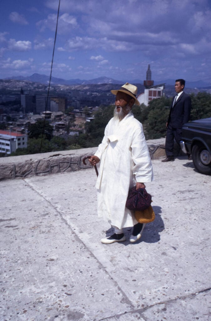 Rear view of a wise grandfather in a traditional Korean Durumagi overcoat with intricate embroidery and pleated skirts, carrying a cane and walking through a historic palace courtyard. This essential Korean style for winter.