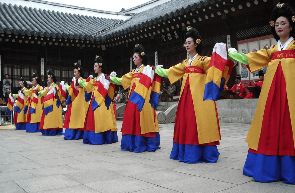 Aesthetic back view of Korean dancers in colorful traditional Hanbok with pleated skirts, performing a traditional Korean dance (Seungmu) in a historic setting.