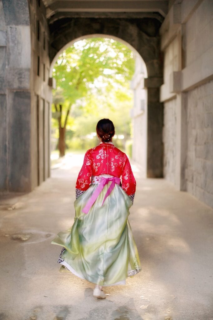 Woman in traditional Korean dress (Hanbok) walking through a Gyeongbokgung Palace courtyard, back view. Essential Hanbok experience for Seoul travel.