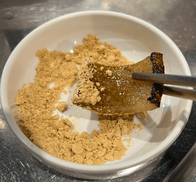 A mouthwatering close-up of a crispy and chewy grilled Korean pork rind (돼지껍데기, Kkeopdegi) being deeply dipped into a mound of nutty, sweet injeolmi (soybean) powder