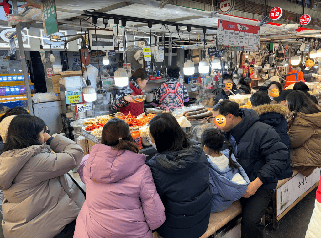 A lively scene inside Gwangjang Market with international tourists sitting at a crowded Pojangmacha (street food stall), enjoying authentic spicy chicken feet (Dakbal) and blood sausage (Sundae).