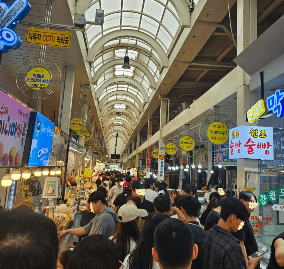 A bustling scene inside the Sokcho Jungang Market, filled with a diverse crowd of locals and tourists exploring the vibrant food alleys.