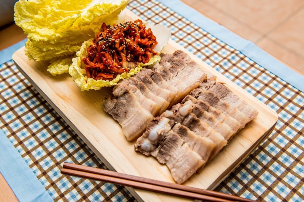 : A traditional Korean Bossam platter featuring succulent boiled pork slices, vibrant red spicy kimchi, and crisp, sweet raw cabbage leaves for wrapping.