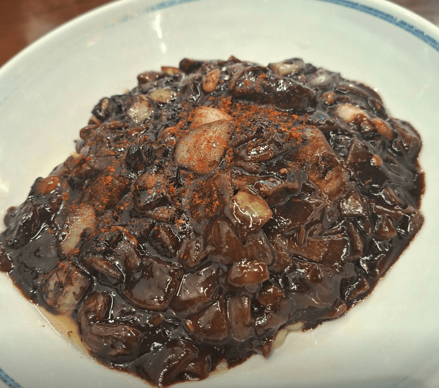 A glossy bowl of Jajangmyeon sprinkled with a dash of red chili powder (Gochugaru), showcasing the authentic Korean way to add a spicy kick and balance the richness of the black bean sauce.