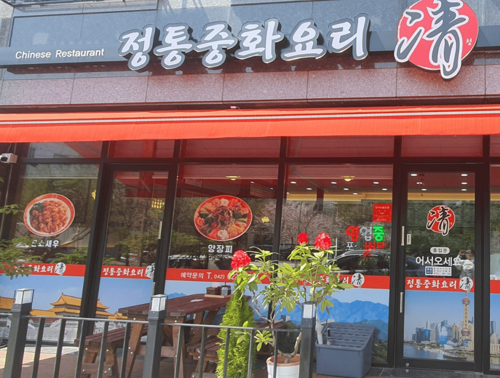 The exterior of a typical neighborhood Korean-Chinese restaurant, featuring a large red sign with gold Hanja characters and a delivery motorcycle parked out front on a cozy street corner.