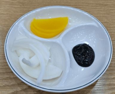 A classic side dish set in a Korean-Chinese restaurant, featuring bright yellow pickled radish (Danmuji), sliced raw onions, and a small portion of black fermented soybean paste (Chunjang).