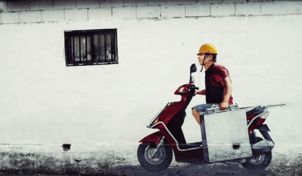 A Korean-Chinese food delivery rider carrying a classic silver steel delivery box, known as 'Chul-ga-bang,' while walking toward a residential building to deliver a fresh meal.