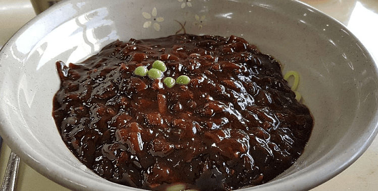A glossy bowl of Korean Jajangmyeon, featuring chewy noodles topped with thick black bean sauce, diced pork, and fresh cucumber garnishes on a traditional restaurant table.