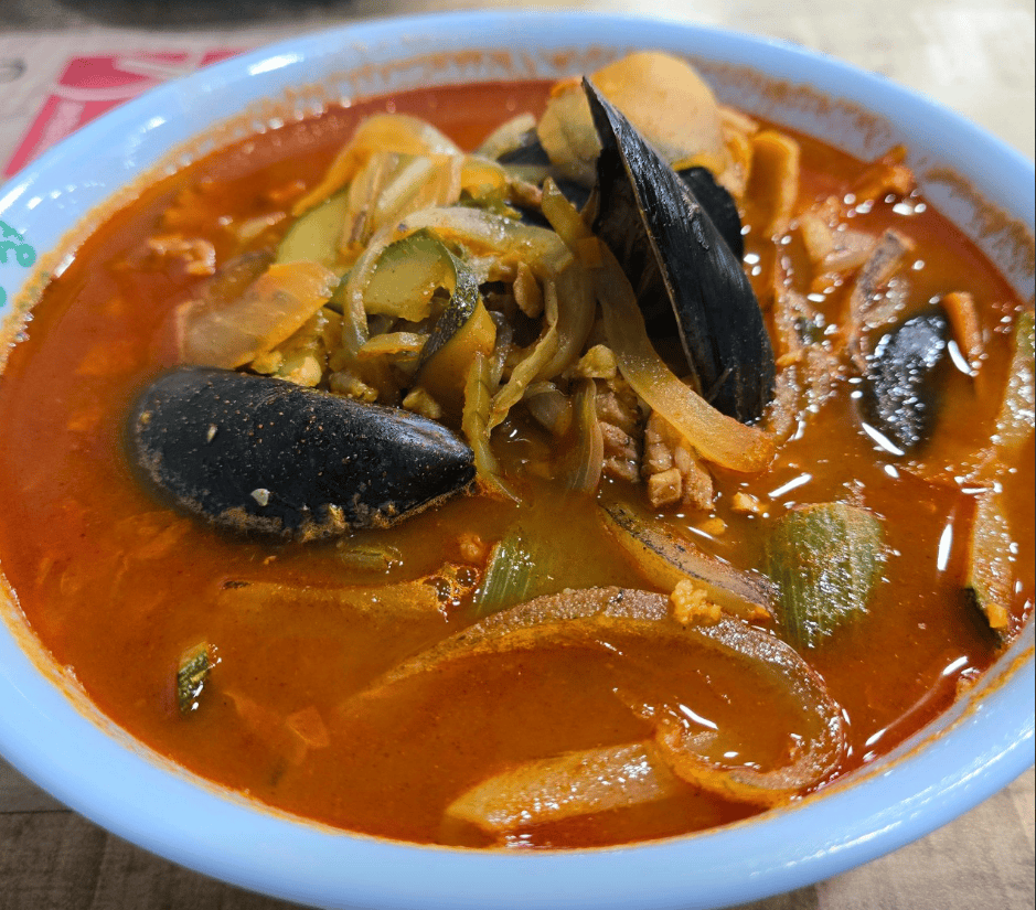 A vibrant bowl of Korean-style spicy seafood noodle soup (Jjamppong), featuring a fiery red broth packed with fresh mussels, squid, and stir-fried vegetables.