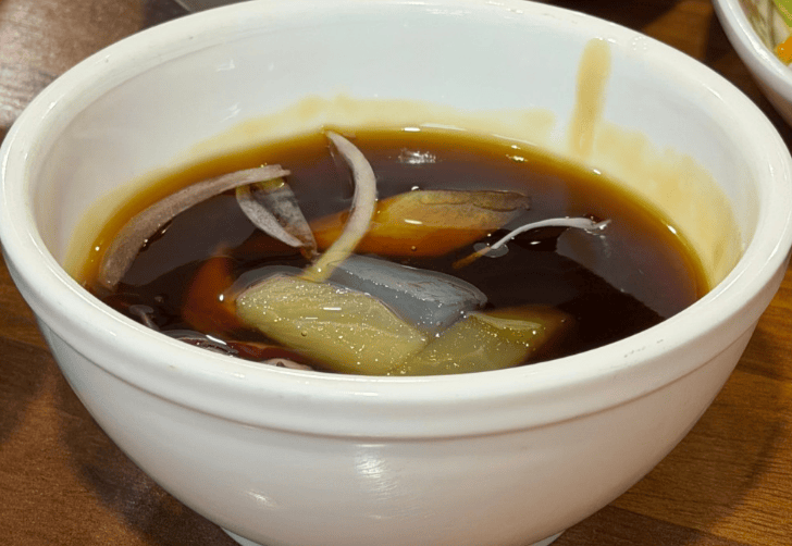 A bowl of glossy, translucent sweet and sour sauce for Tangsuyuk, featuring chunks of yellow pineapple, orange carrots, and crisp white onions.