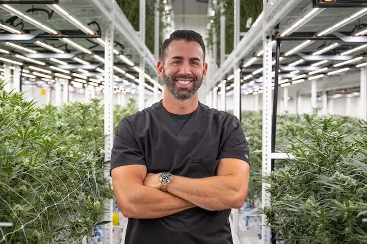 1. Man in black scrubs inspects commercial cannabis under LED grow lights. 2. Cannabis cultivation expert with arms crossed amid LED-lit cannabis plants. 3. Grower in black scrubs stands by cannabis under advanced LED grow lights. 4. Worker oversees cannabis plants illuminated by modern cultivation lighting tech. 5. Cannabis lighting science: Expert monitors healthy plants under LED grow lights.