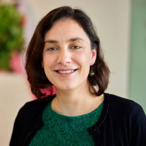 Une femme souriante aux cheveux bruns mi-longs, vêtue d'une chemise verte et d'un gilet noir, représentant les solutions d'éclairage LED Fluence pour la culture du cannabis, se tient à l'intérieur sur une toile de fond floue, mettant en évidence une production végétale optimisée dans des environnements contrôlés. width= (en anglais)