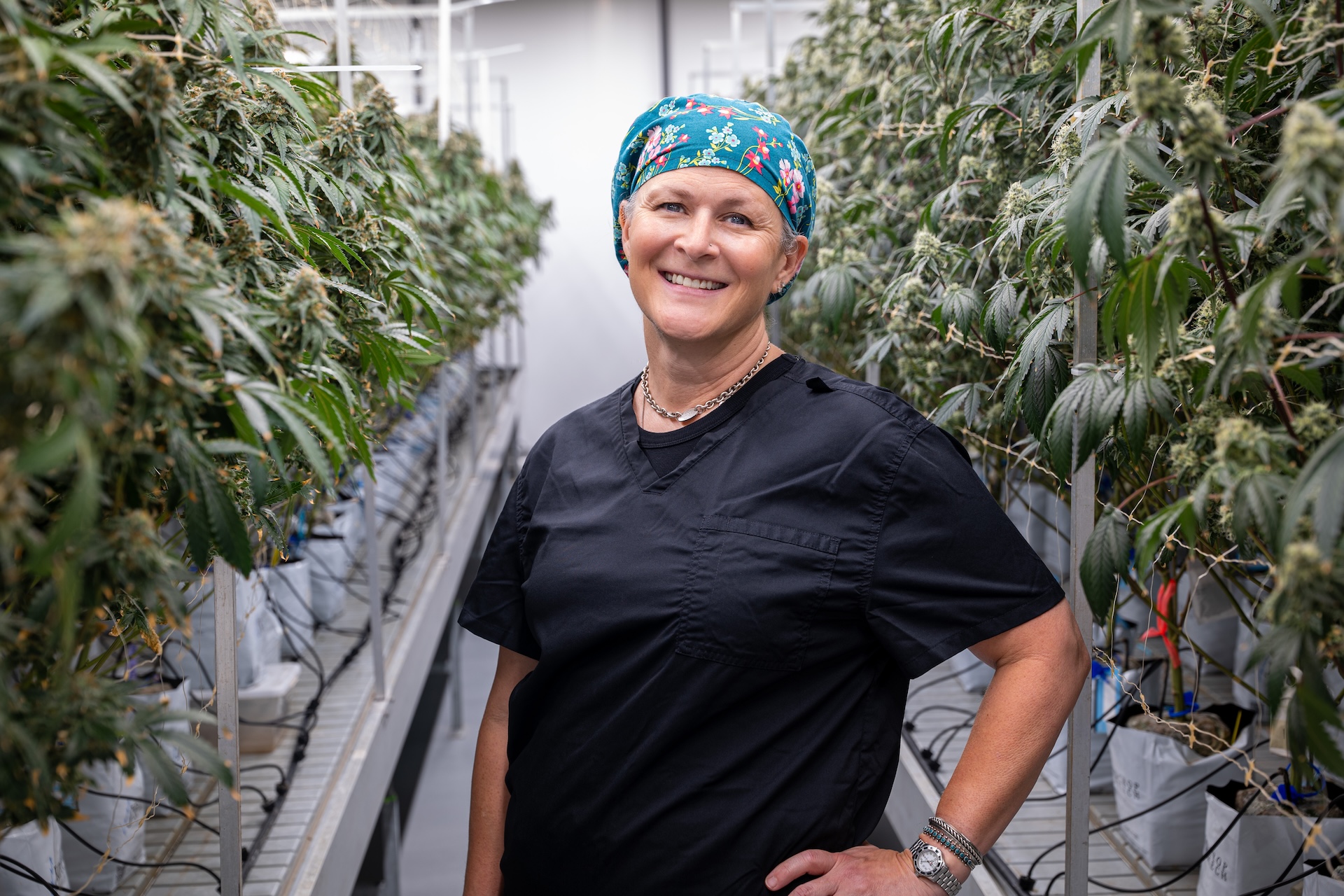 Person in scrubs under LED grow lights smiles in a commercial cannabis cultivation facility.
