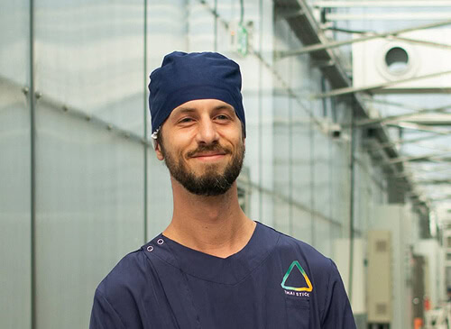 A man in blue scrubs stands beneath LED grow lights for cannabis cultivation, holding a Thai Stick.