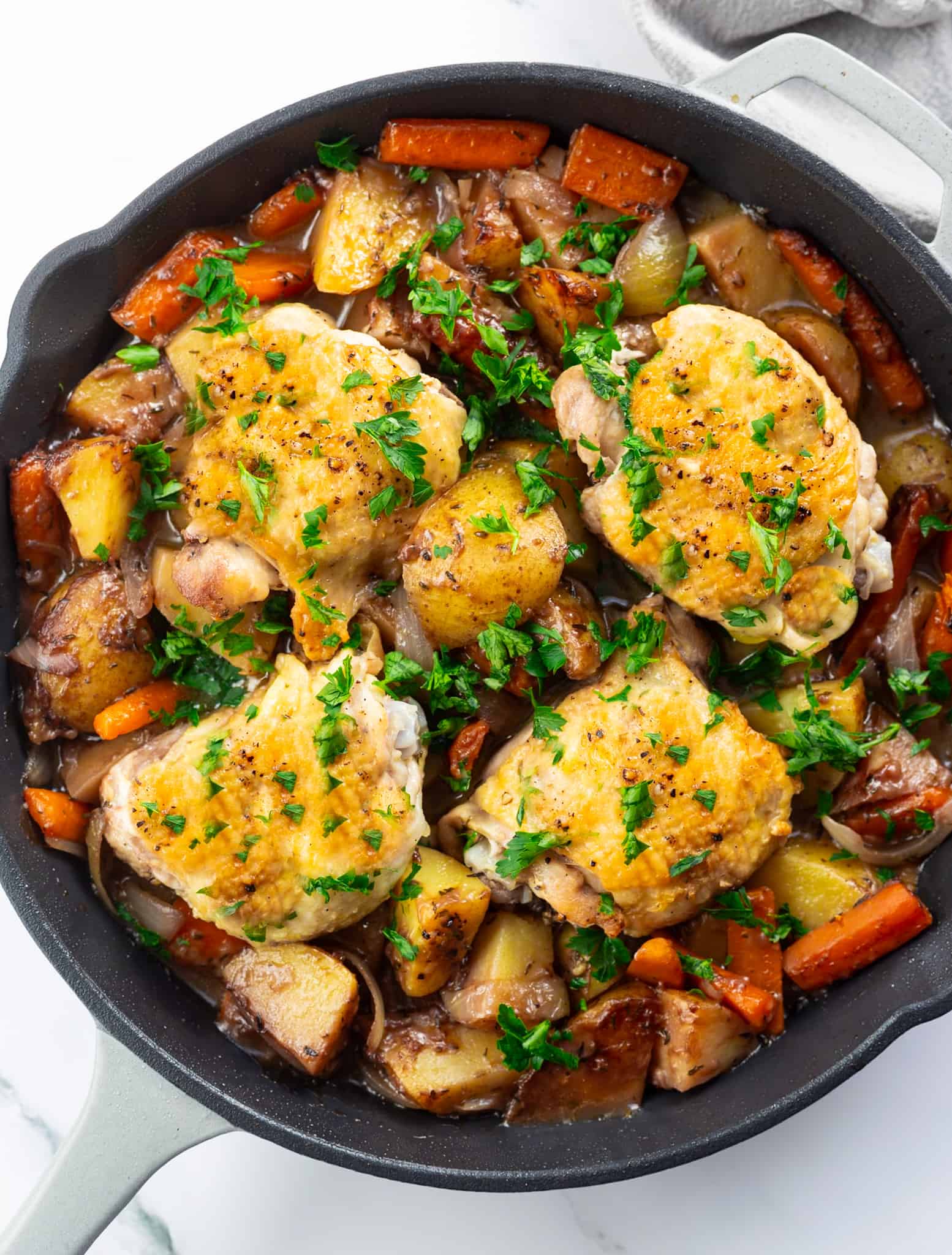 A cast iron pan of braised chicken thighs surrounded by cooked potatoes, carrots, and onions and garnished with fresh parsley.