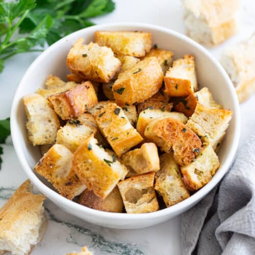 A bowl of homemade croutons surrounded by torn bread and a bushel of fresh parsley.