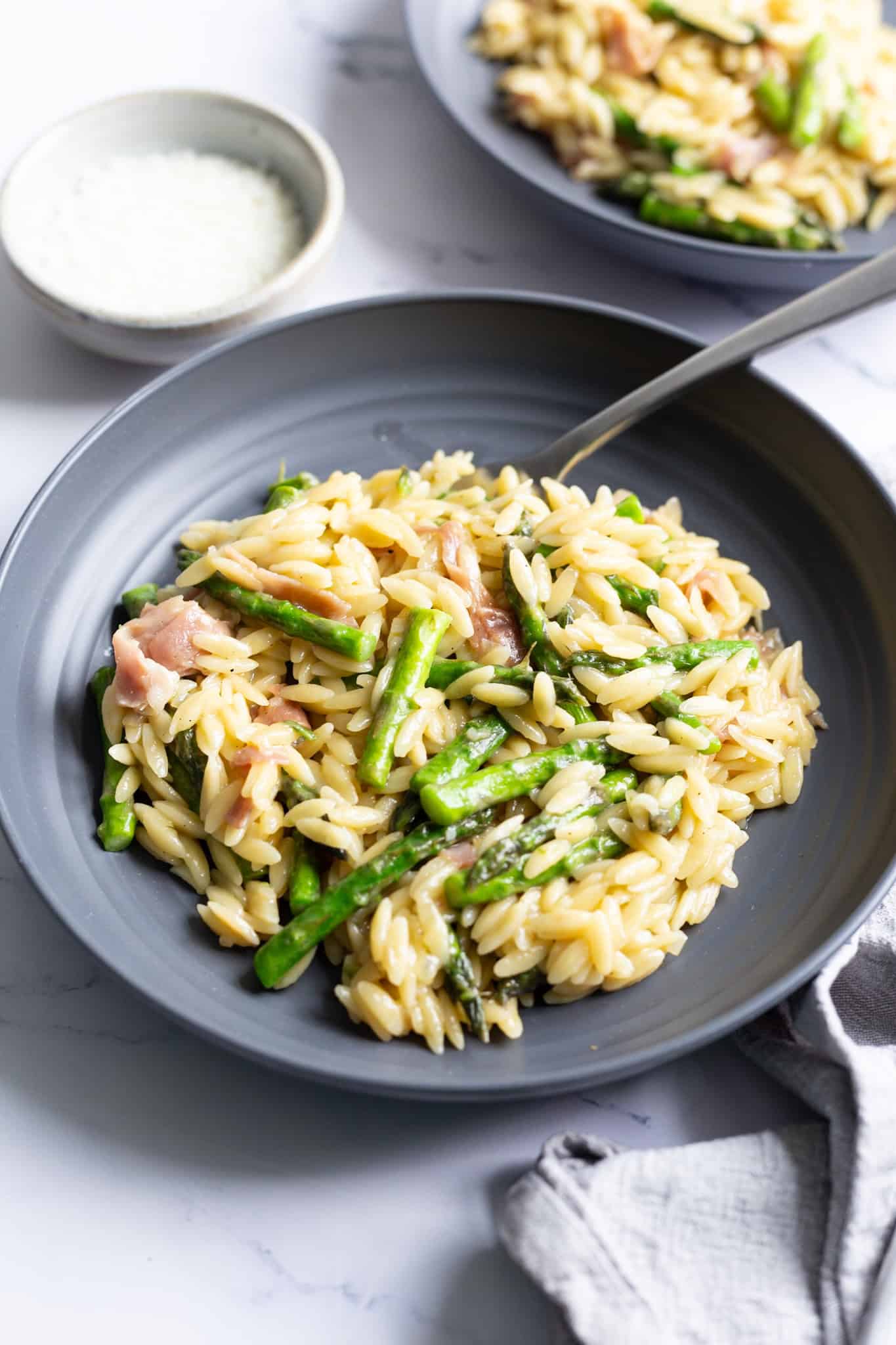 A plate of orzo pasta with asparagus and prosciutto, with a small bowl of grated Parmesan on the side.