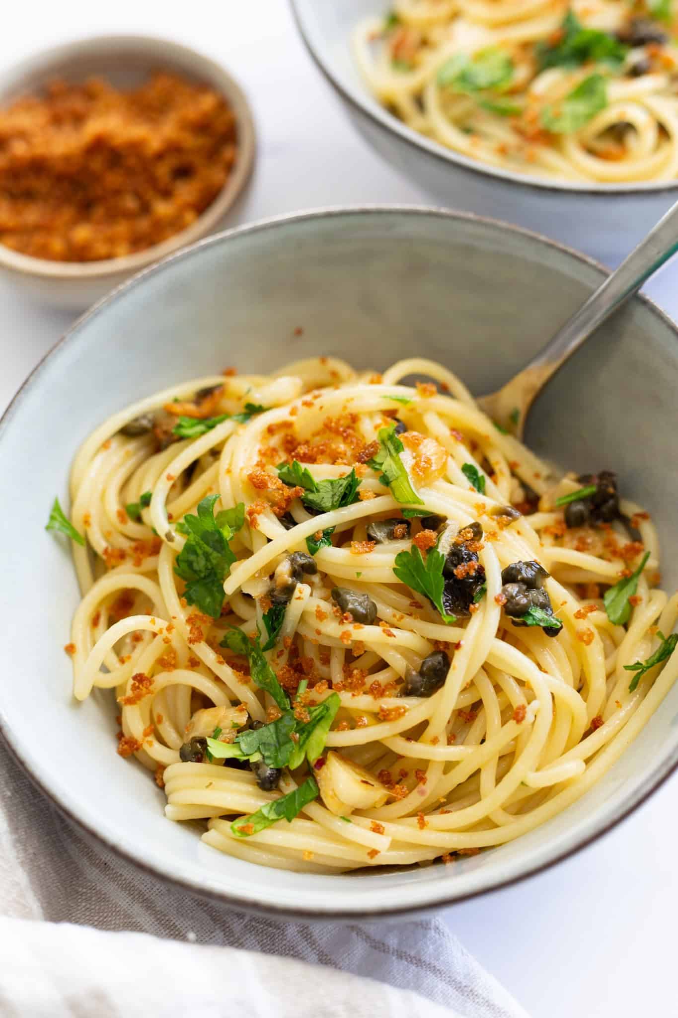 A light grey bowl of spaghetti with lemon, capers, and roasted garlic.