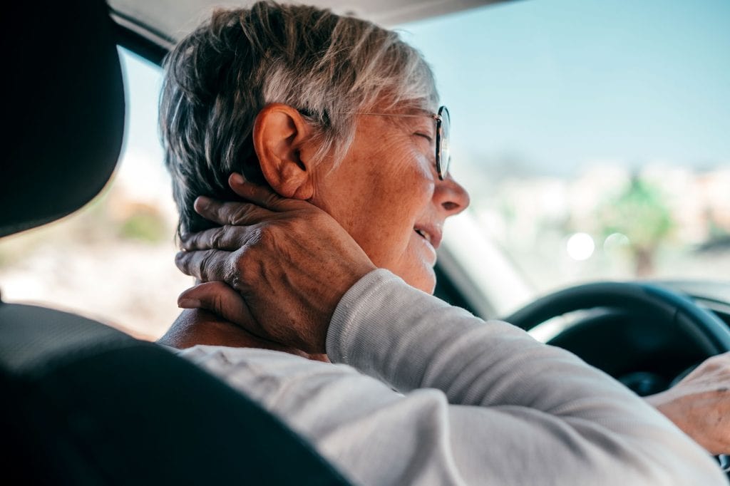 Elderly woman experiencing neck pain while driving, seeking healthcare support.