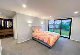 Bright modern bedroom featuring a large bed with colourful bedding, walk-in closet, and sliding glass doors overlooking a green outdoor space.