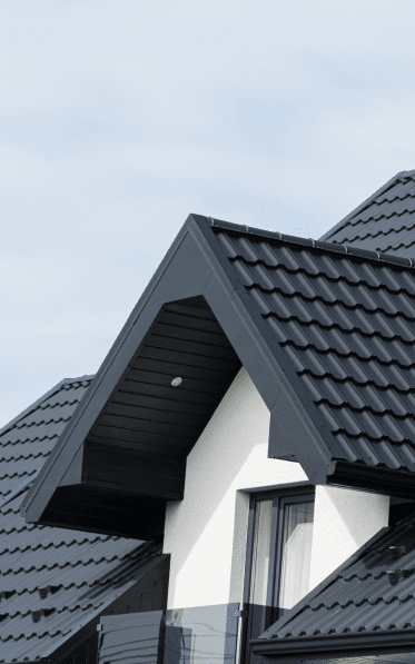 A close-up architectural photograph of a modern residential roof with dark gray metal shingles. The image shows a prominent roof peak with a black soffit and fascia board. The roof has clean, straight lines and sharp angles. A white-painted wall section is visible beneath the roof overhang, creating a strong contrast with the dark roof material. A large rectangular window with black trim is partially visible in the frame. The composition is shot against a light blue sky with wispy white clouds. The metal roofing tiles are arranged in overlapping rows with a distinctive wave pattern. The image has a minimalist, contemporary aesthetic with strong geometric shapes and clean lines.