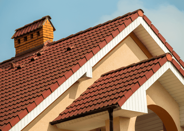 A close-up architectural photograph of a residential roof with terracotta-colored clay tiles arranged in a uniform pattern. The roof has a prominent brick chimney in orange-brown color positioned on the left side. The roofline features white trim boards along the eaves, creating clean geometric lines against the warm-colored tiles. A decorative architectural detail shows a peaked gable with white trim and a small overhanging roof section. The background shows a clear blue sky. The roof tiles are in excellent condition, showing a rich reddish-brown color with subtle variations in tone. The composition is shot at a slight angle, emphasizing the architectural details and craftsmanship of the roof construction.