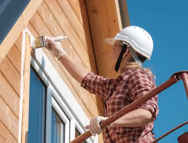 A close-up photograph of a caucasian woman painting the exterior of a wooden house on a sunny day. The worker wears a white hard hat, a red and black plaid flannel shirt, and white protective gloves. She is painting white trim around a window opening using a long-handled paint brush. The wooden siding of the house is natural pine-colored cedar planks. A blue sky is visible in the background. The worker is positioned on a ladder, with the ladder partially visible in the frame. The lighting is bright and natural, creating distinct shadows on the wooden siding. The image is shot from a slight low angle, emphasizing the height of the work being performed.