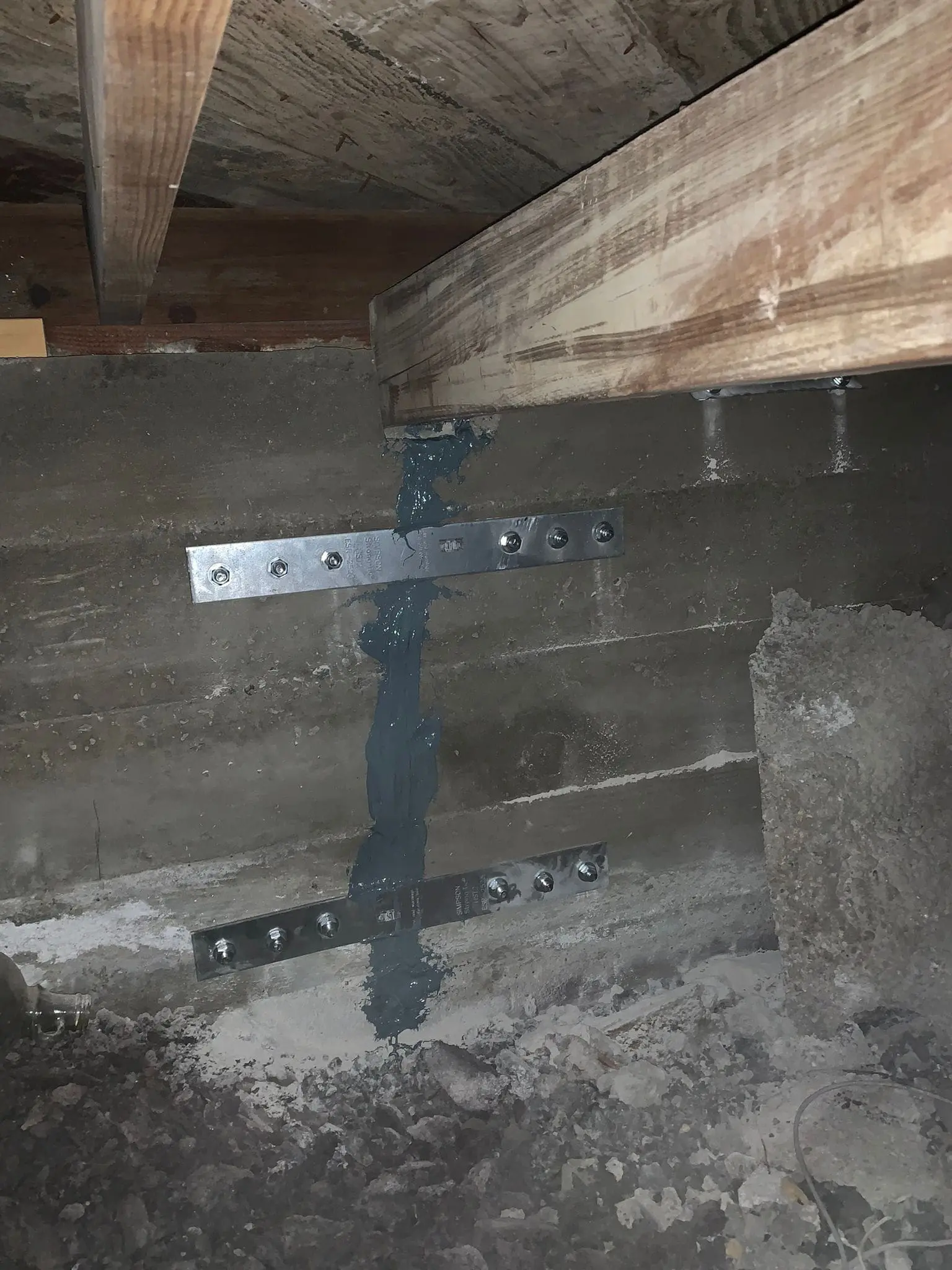 A close-up photograph of a concrete foundation wall with visible water damage. The wall has a horizontal metal strap with multiple bolt holes mounted to it. A blue-gray water stain runs vertically up the concrete wall, starting from the metal strap. The concrete wall shows signs of aging with rough, weathered texture and patches of exposed aggregate. At the top of the frame, there is a wooden beam partially visible. The concrete floor at the bottom of the frame is deteriorating with exposed gravel and debris. The lighting in the image is dim and creates shadows in the corners of the concrete structure. The metal strap is a standard structural bracket with multiple circular mounting holes arranged in a line.