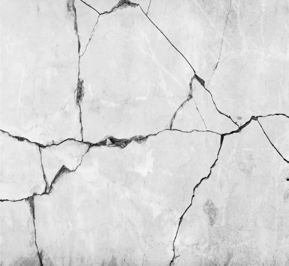A high-contrast black and white photograph of a cracked concrete wall. The wall features a complex network of diagonal and curved cracks that divide the surface into irregular geometric sections. The cracks are dark gray against the light gray concrete background. The texture of the concrete is rough and weathered, with subtle variations in the gray tones. The cracks create a pattern of angular fragments, with some sections larger than others. The image has a monochromatic quality with strong contrast between the light and dark areas. The composition fills the entire frame with the concrete surface, showing the architectural detail of the wall's surface damage and weathering.