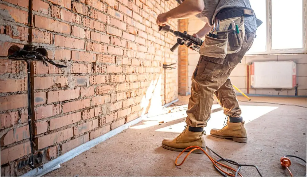A construction worker in khaki work pants and tan leather work boots stands in a partially renovated room with exposed red brick walls. The worker is holding a power tool and is mid-motion, with tools visible in their utility belt. The room has a concrete floor and natural sunlight streams in from a window on the right side, creating a bright spot on the floor. Construction materials and tools are scattered on the floor, including an orange extension cord. The brick wall shows signs of wear with visible mortar lines and weathering. The image is cropped to show the worker from mid-thigh down, with the focus on the practical workwear and tools. The lighting creates strong shadows and highlights the texture of both the brick wall and the work boots.