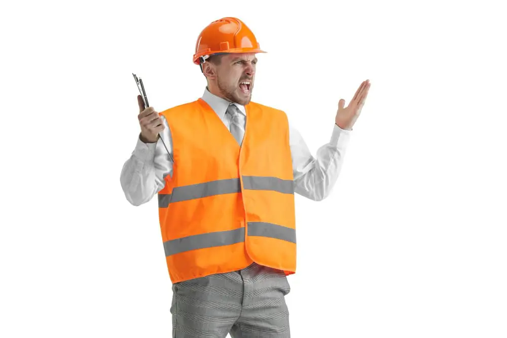 A professional photograph of a caucasian man construction worker in a bright orange safety vest with reflective silver strips, wearing a white dress shirt and gray tie. The worker is wearing a bright orange hard hat and is captured in an expressive pose with his mouth open in an angry shout. He is holding a walkie-talkie in his right hand while his left arm is raised with an open palm gesture. The worker is wearing gray patterned pants. The background is pure white, creating a stark contrast with the orange safety equipment. The image is a full-body shot, showing the worker from head to toe. The lighting is even and professional, typical of studio photography. The worker's expression conveys frustration or anger, with furrowed brows and tightened facial muscles.