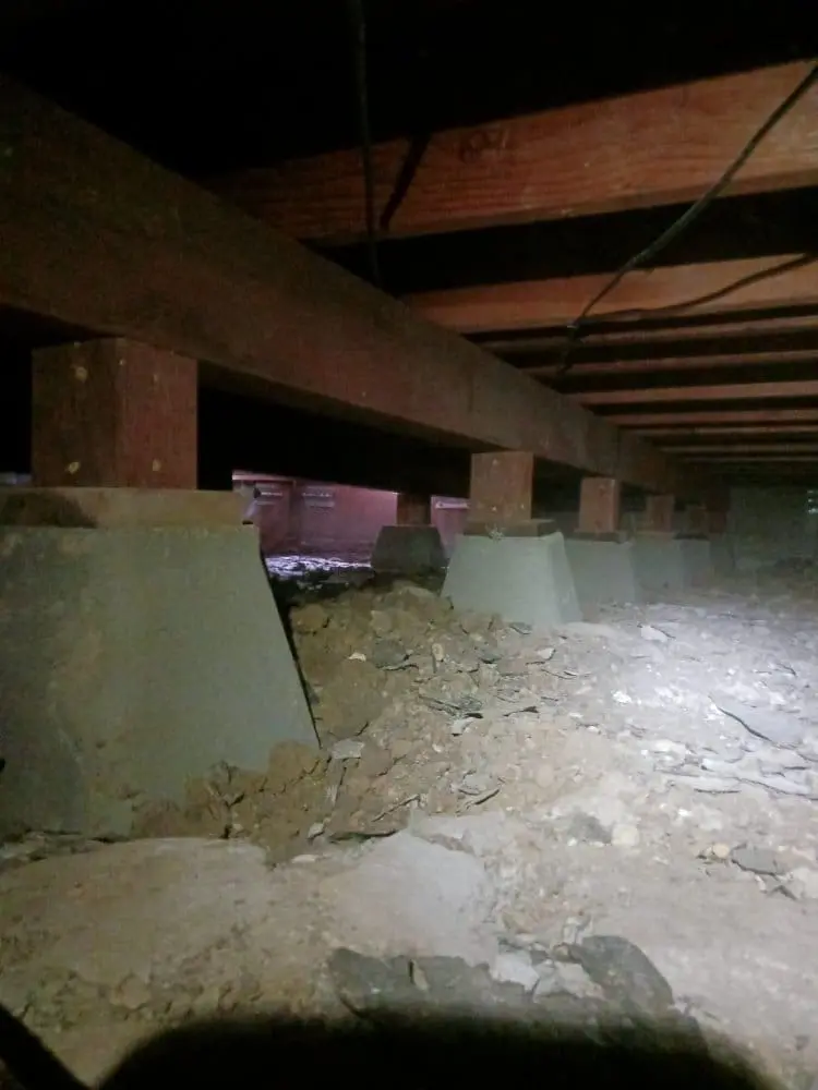 A dimly lit basement crawlspace with exposed wooden support beams and concrete pillars. The concrete pillars are painted in a light sage green color and are supported by concrete bases. The floor is made of concrete with visible cracks and deterioration. The wooden beams above are weathered and show natural wood grain patterns. The lighting is harsh and creates strong shadows, casting a reddish tint in some areas. The space is industrial and unfinished, with exposed utilities and pipes running along the wooden beams. The ceiling is low, creating a confined feeling in the space. The concrete pillars are arranged in a regular pattern, supporting the weight of the structure above. The overall image has a moody, underground atmosphere with strong contrast between the light and shadow areas.