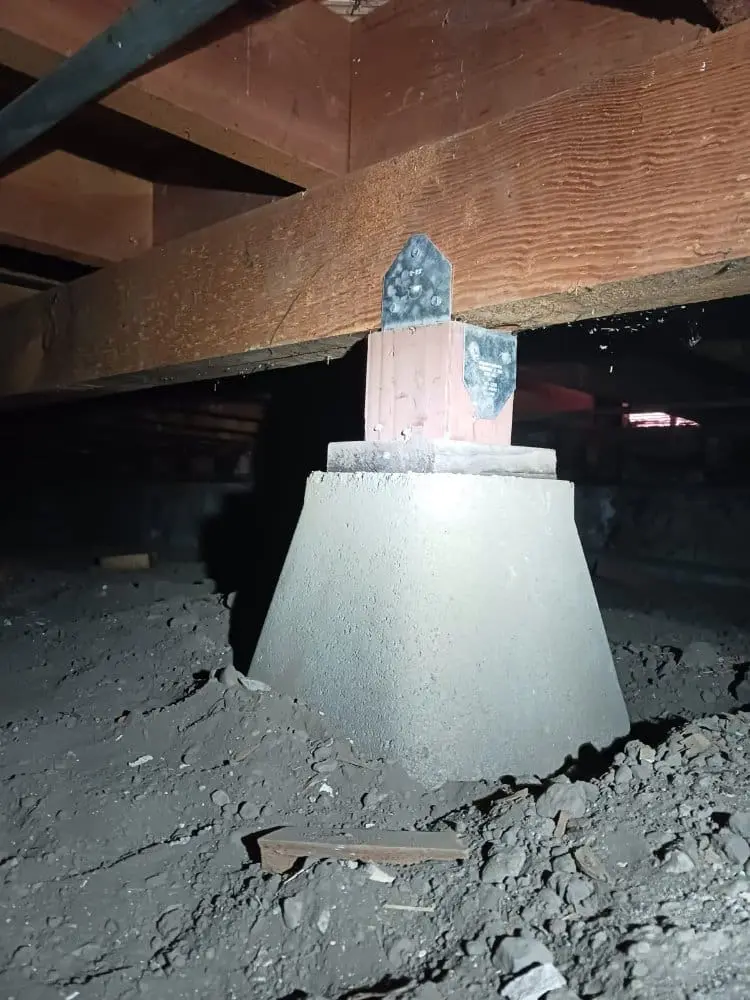 A close-up photograph of a concrete support column foundation in a dark crawl space. The column has a light gray, conical-shaped base that tapers upward. At the top of the column is a small, weathered concrete marker with blue-tinted markings. The surrounding area shows exposed dirt and debris on the ground, with scattered rocks and construction materials. Above the column, there are wooden support beams visible, showing natural wood grain patterns and a reddish-brown color. The lighting is dim and creates shadows in the dark space, giving the image a moody, industrial atmosphere. The concrete column is part of a structural support system, with its distinctive conical shape designed to distribute weight evenly.