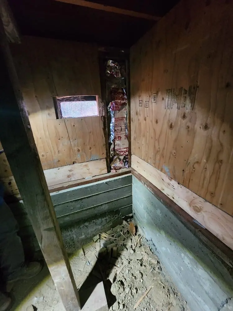 A deteriorating interior room with plywood walls and a concrete floor. The walls are covered in unfinished plywood panels with visible wood grain and knots. A small rectangular window is visible on the upper left wall, letting in natural light. The concrete floor is covered in debris, dirt, and scattered construction materials. The walls meet at a right angle, creating a corner space. The lighting creates shadows in the corners and along the walls. The plywood shows signs of weathering and aging, with some areas darker in color. The concrete foundation shows signs of moisture damage with a greenish tint in the lower portion. The overall image has a dark, abandoned atmosphere with natural lighting creating contrast in the space.