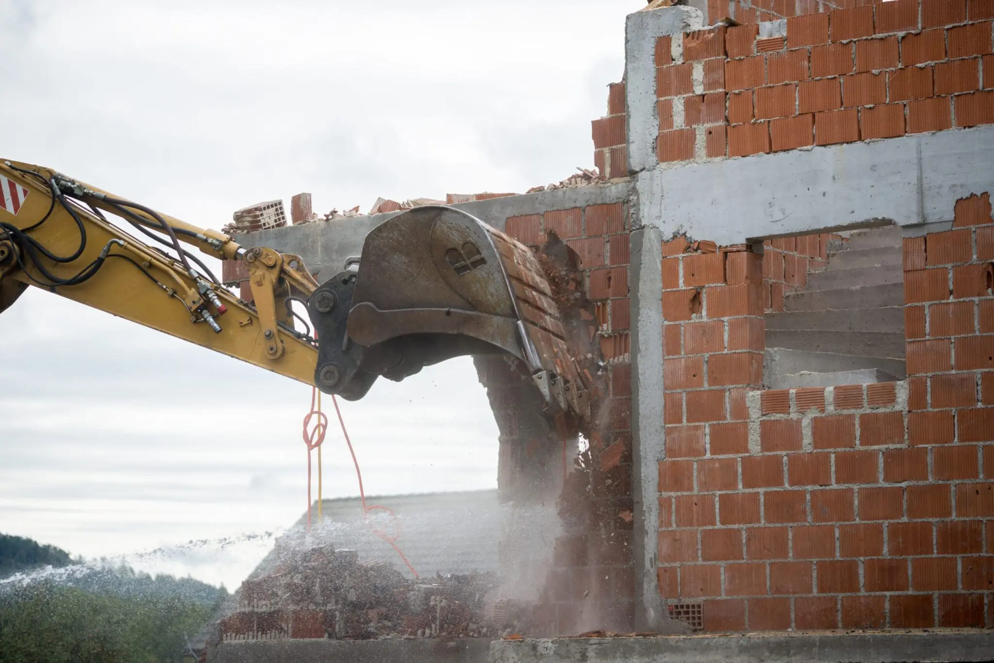 A demolition scene showing a yellow hydraulic excavator arm with a large shearing tool tearing through a red brick wall. The brick wall is partially demolished, with concrete and brick debris flying into the air. The wall is constructed with standard orange-red bricks in a traditional pattern, with concrete blocks visible at the top and bottom. The excavator's metal shears are cutting through the brick, creating a dramatic tearing effect. Dust and debris are being sprayed into the air from the point of demolition. The background shows a cloudy gray sky and a distant tree line. The image captures the industrial process of building demolition in progress, with the contrast between the hard, angular machinery and the traditional brick construction.