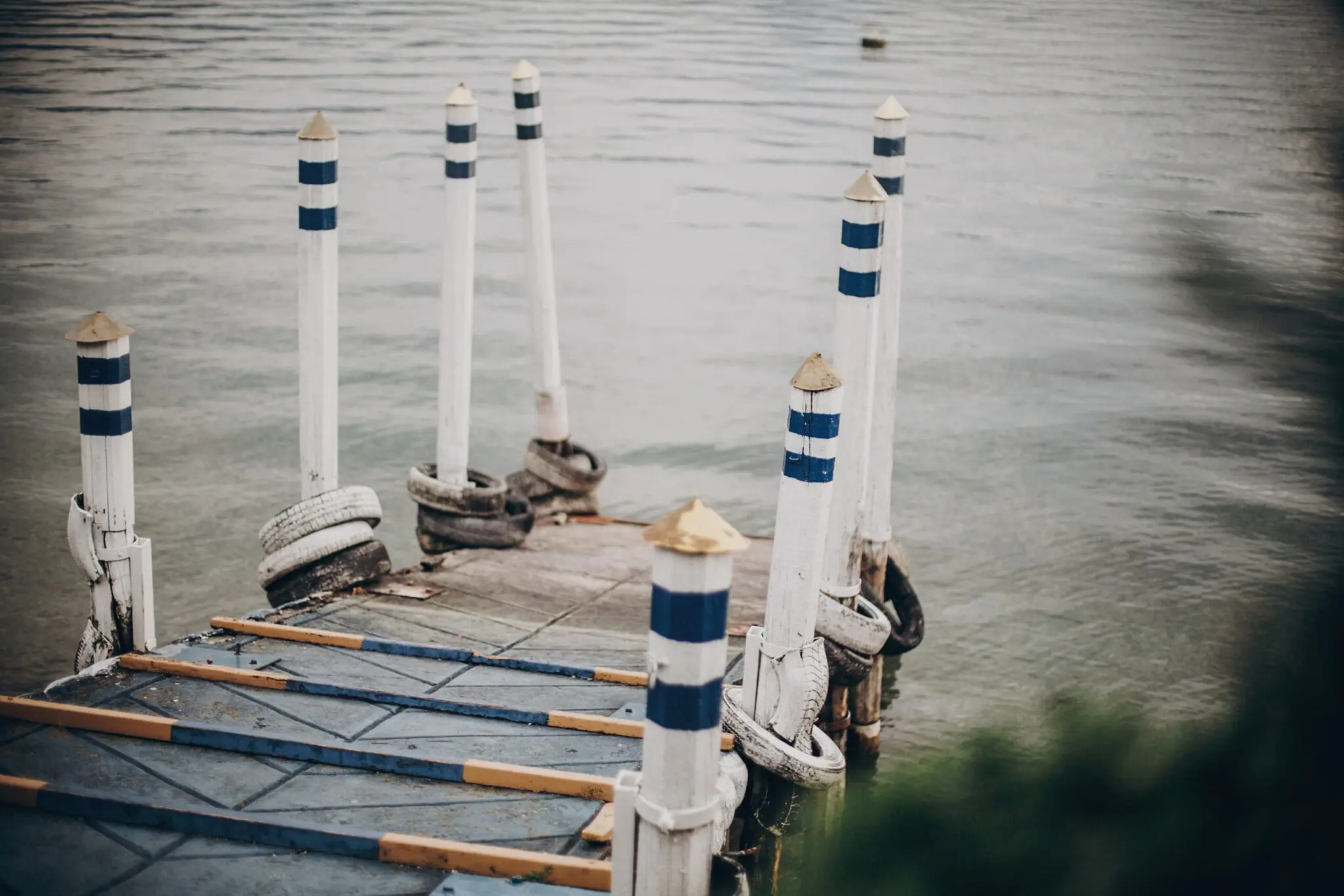 A moody photograph of a weathered dock with white and navy blue striped bollards against a gray water background. The dock is constructed of concrete with wooden planks and metal supports. The bollards are evenly spaced and feature a distinctive striped pattern of white and navy blue bands, topped with golden caps. The water in the background has a smooth, rippled texture and a muted gray-blue color. The image has a vintage, desaturated quality with a slight vignette effect around the edges. In the foreground, there are wooden support beams and metal fixtures attached to the dock's structure. The composition is shot from a slightly elevated angle, creating depth and leading lines toward the water. The overall atmosphere is moody and nautical, with the striped bollards creating a strong visual pattern against the monochromatic water backdrop.