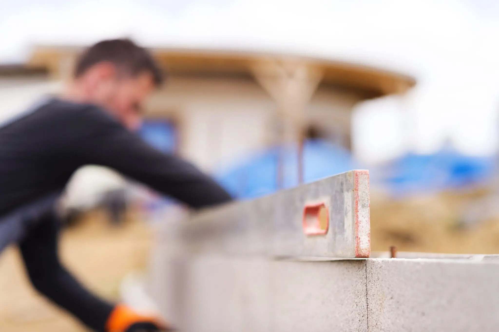 A close-up photograph of a construction worker using a level tool on a concrete block wall. The worker is wearing a black long-sleeved shirt and is partially visible in the blurred background. The level tool is gray with red markings and is positioned horizontally against the concrete block. The concrete blocks are light gray in color with visible seams between them. The image has a shallow depth of field, creating a soft blur effect in the background where there is a wooden structure with a brown roof. The lighting is natural and even, creating subtle shadows on the concrete surface. The composition is shot at a slight angle, with the level tool positioned diagonally across the frame.