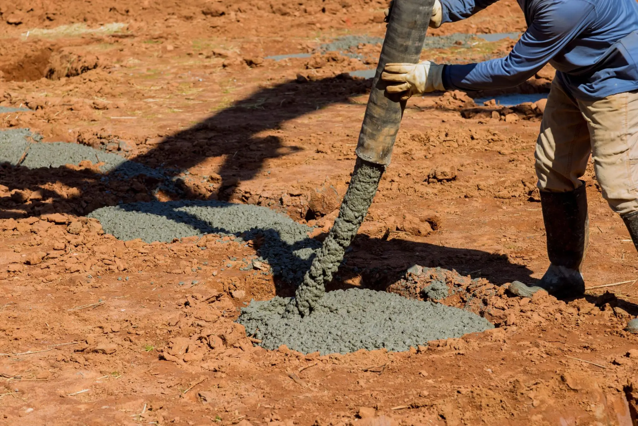 A close-up photograph of a construction worker pouring gray concrete onto reddish-brown dirt ground. The worker is wearing a blue long-sleeve shirt, khaki pants, and black rubber boots. They are holding a black metal pipe from which the concrete is flowing in a steady stream. The concrete forms a curved, puddle-like shape on the ground. The dirt surface has a rough, granular texture and is exposed earth. The lighting is bright and natural, casting a distinct shadow of the worker and equipment on the ground. The image is shot from a low angle, focusing on the pouring action and the immediate ground surface.