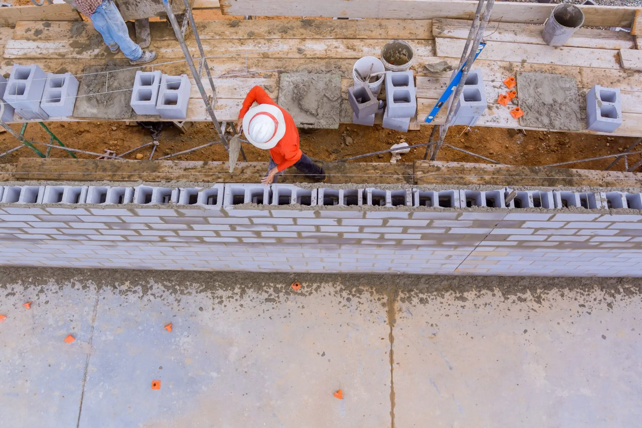 An aerial construction site photograph showing a worker in a bright orange safety vest and white hard hat laying concrete blocks. The worker is positioned on wooden scaffolding, working on a wall foundation. Multiple gray concrete blocks are arranged in a row, with some blocks already laid in place forming a wall base. The wooden scaffolding is made of light-colored planks. Orange safety cones are scattered on the ground near the construction area. The image is taken from directly above, showing the precise layout of the construction work in progress. The background shows exposed dirt and construction materials. The lighting is natural daylight, creating clear visibility of all construction elements.