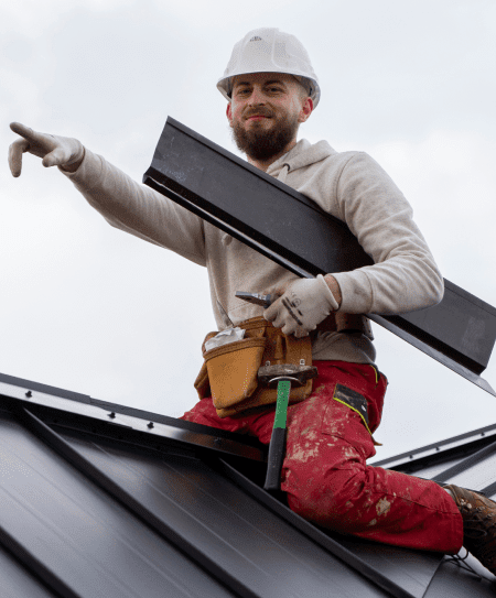 Smiling roofer in a white hard hat pointing while holding a metal panel.