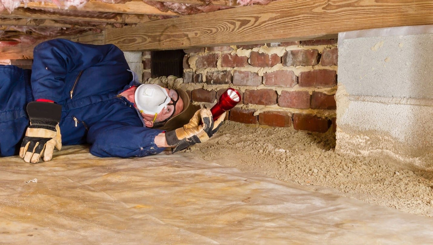 Man in blue coveralls inspecting a crawl space with a flashlight.
