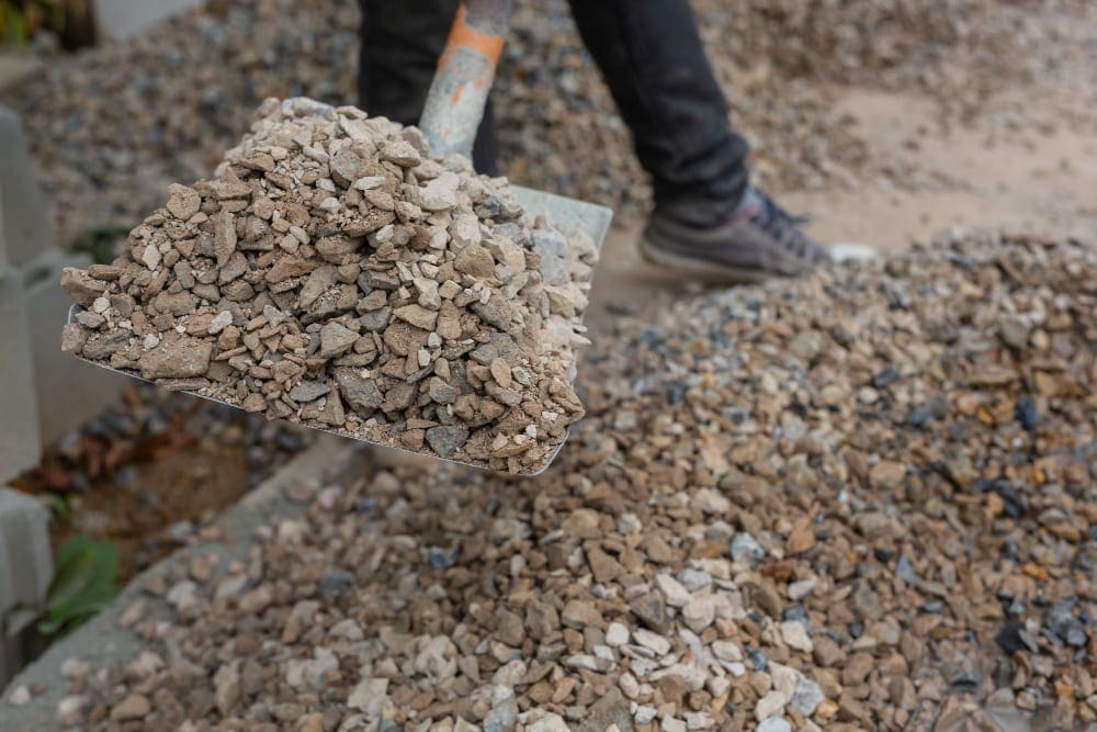 Close-up of a shovel full of gravel with a person's legs in the background.