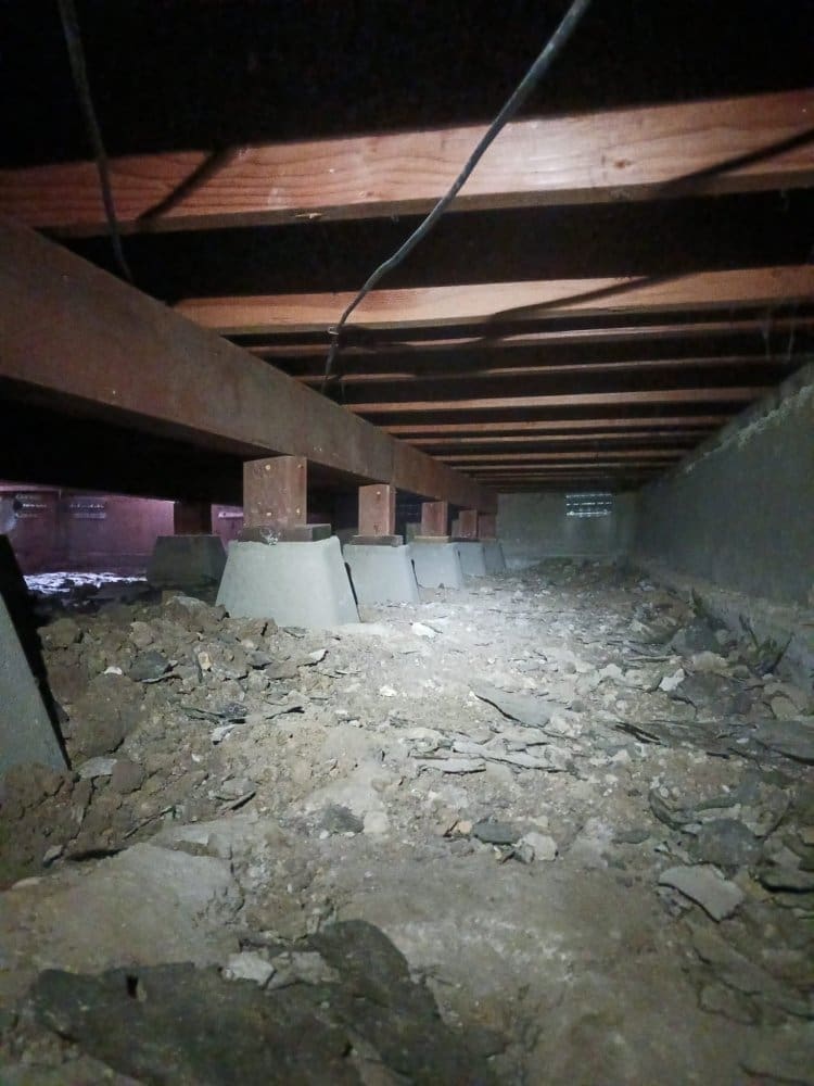 Crawl space with wooden beams, concrete piers, and dirt floor.