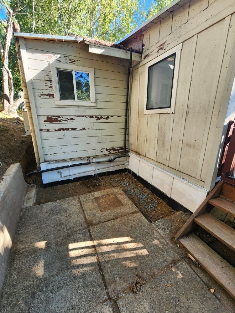 Exterior view of a weathered house with a concrete patio and exposed foundation.