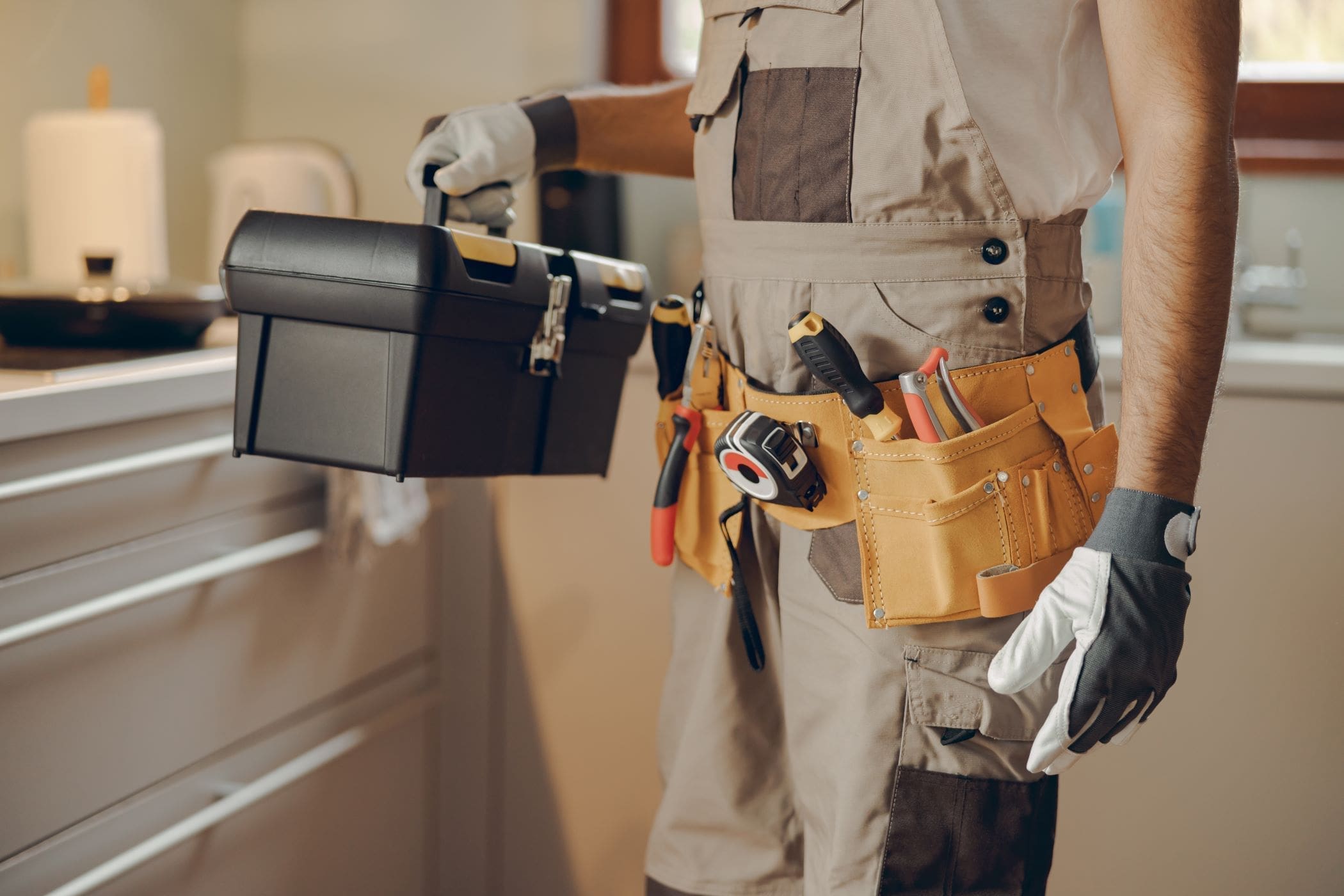 Person in work uniform holding a black toolbox and wearing a tool belt.