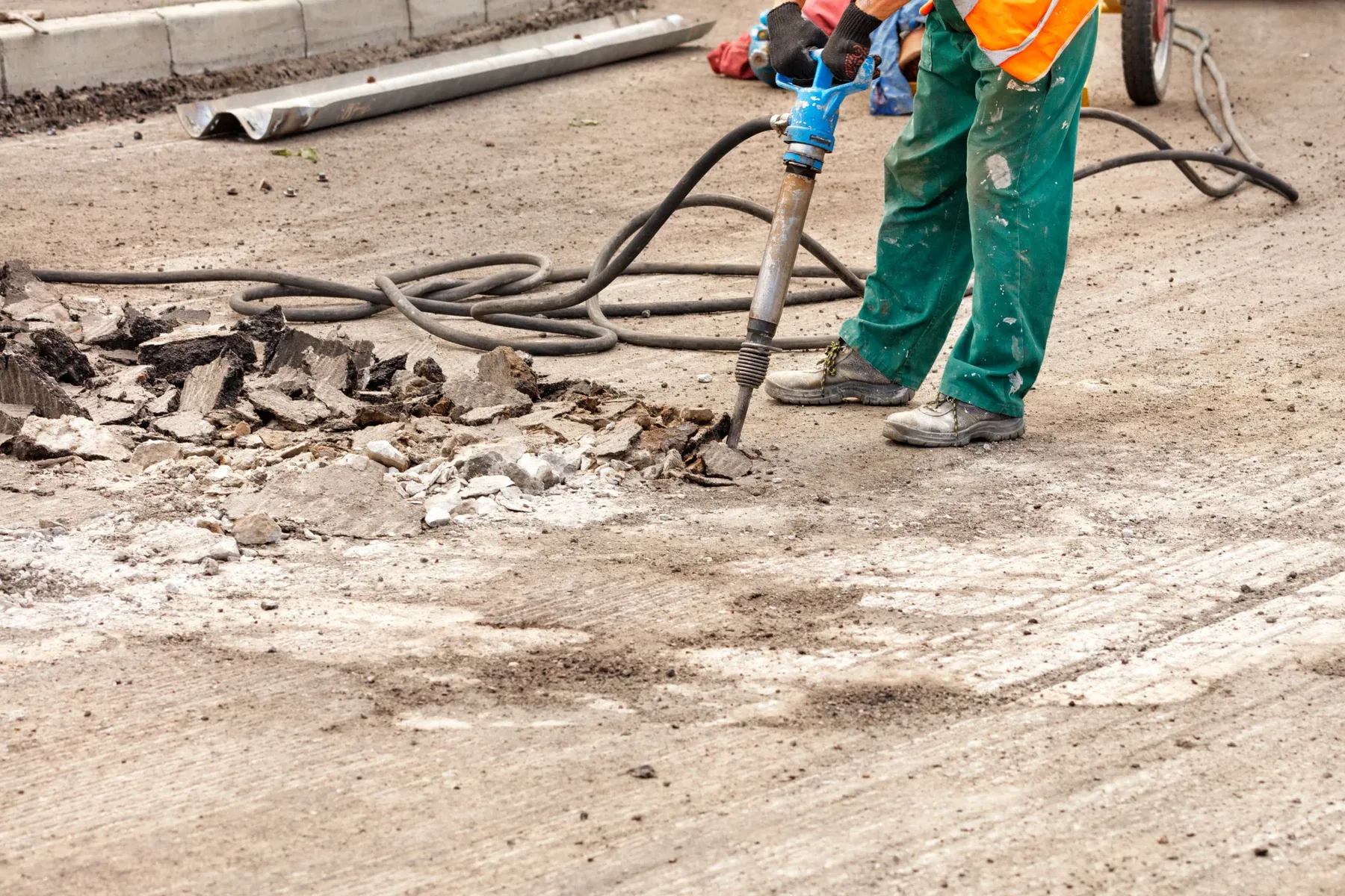 Worker using a jackhammer on asphalt, surrounded by debris and tools.