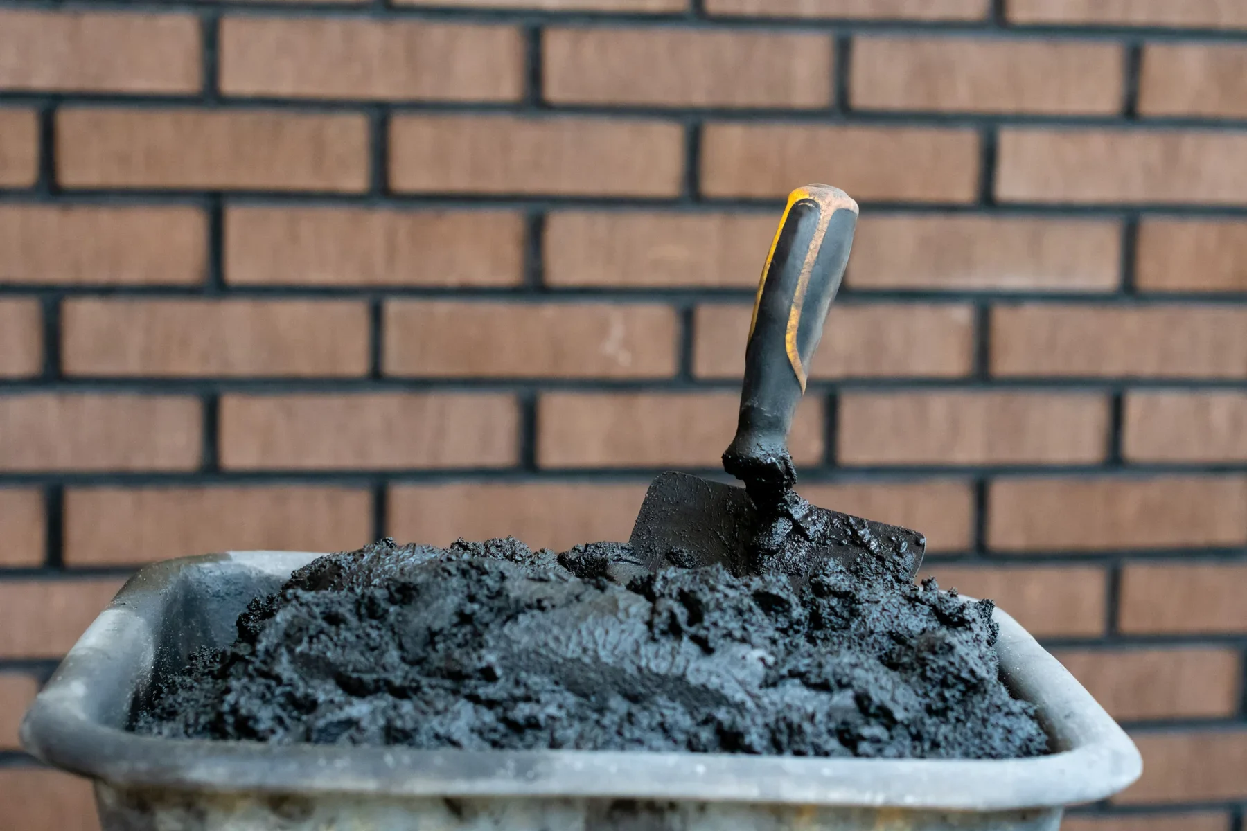 Black shovel in a bucket of dark soil against a brick wall.